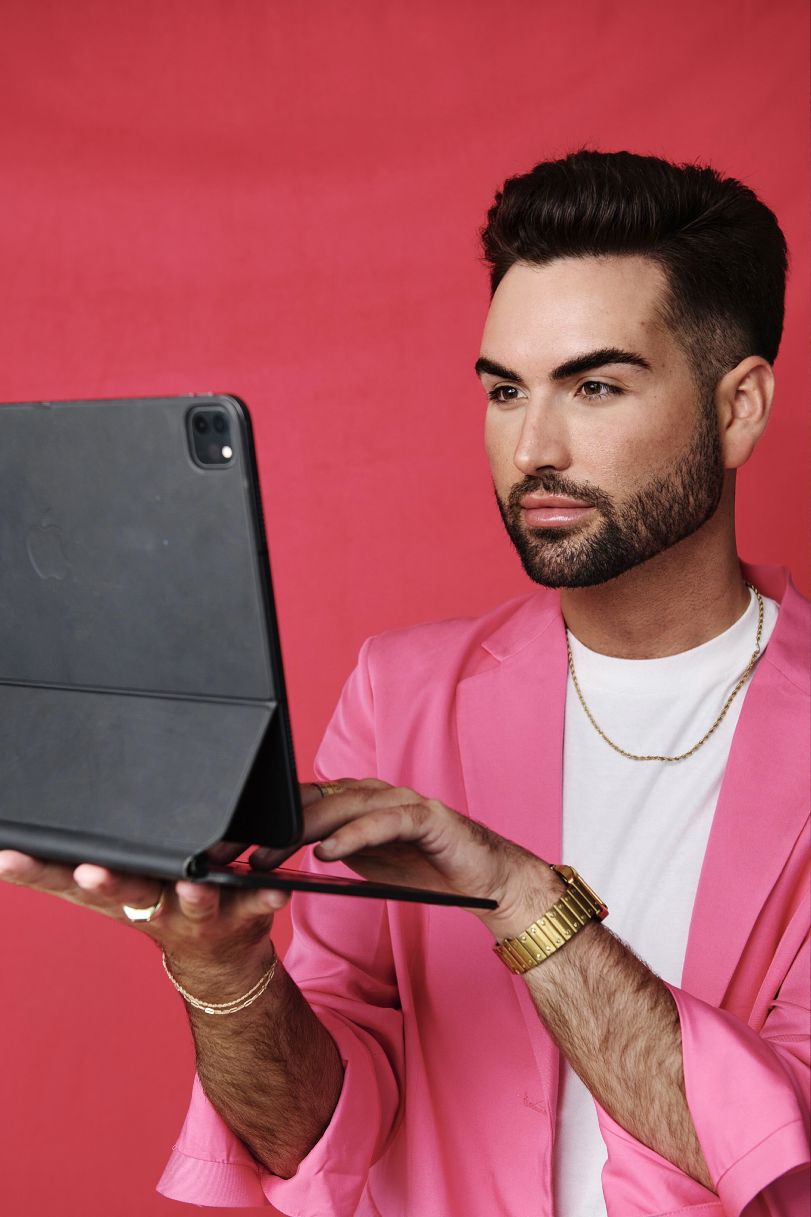 young dark-haired man in a white tee and pink blazer holding up an ipad against a deep fuscia backdrop.
